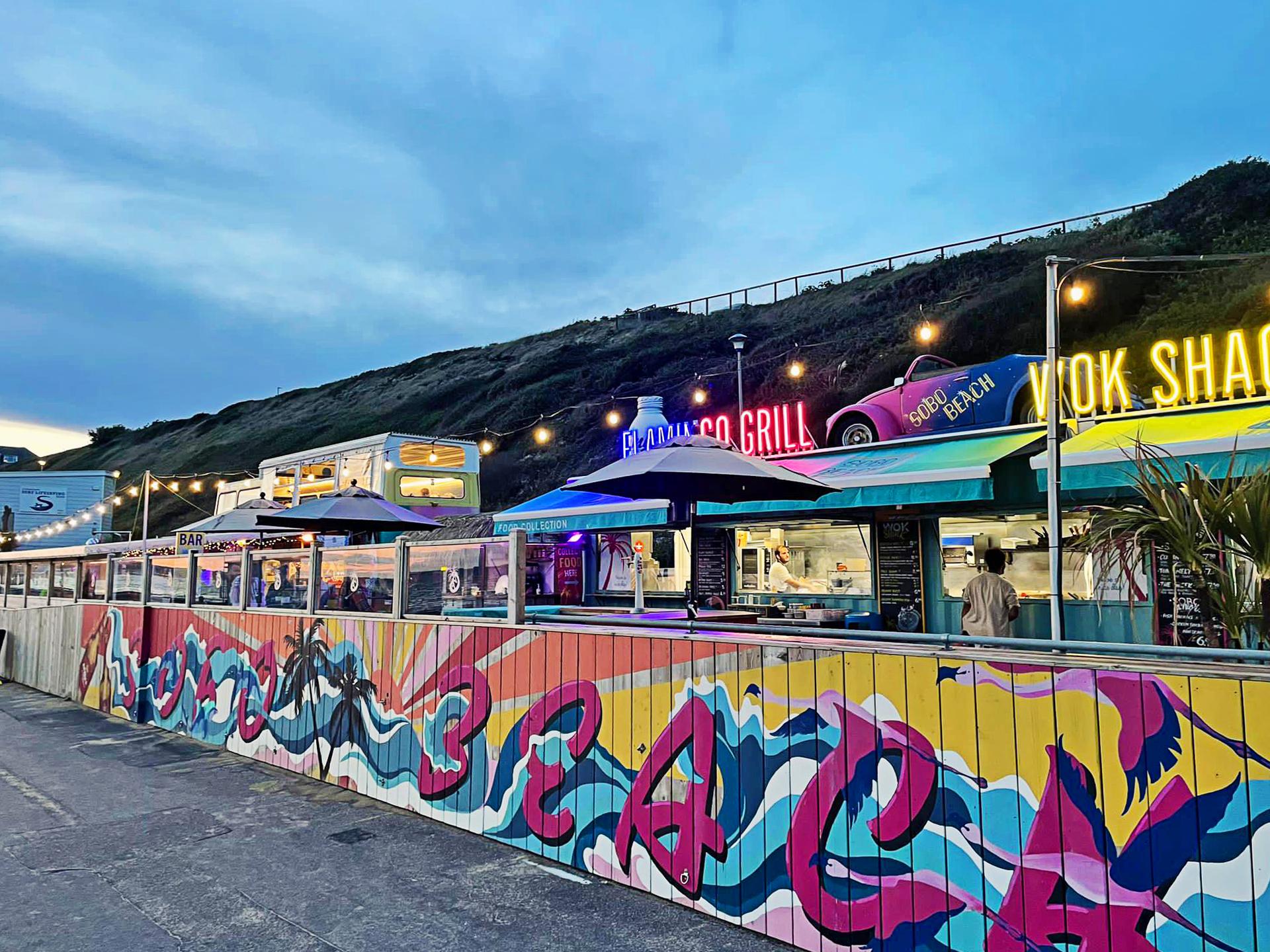 SOBO Beach at dusk: neon Flamingo Grill and Wok Shack signs, the pink Beetle, festoon lights and the hand-painted flamingo and wave mural
