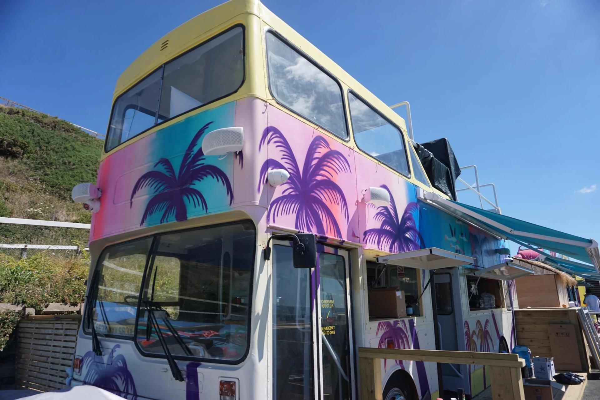 SOBO Beach’s vintage double-decker bus, hand-painted with pink, teal and purple airbrushed palm trees under a blue sky